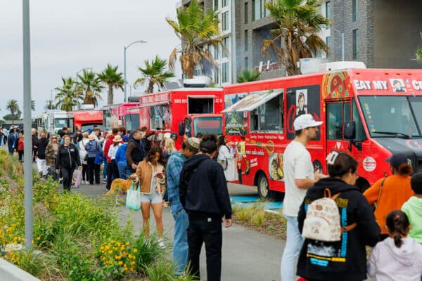 Crowd lined up at colorful food trucks along a waterfront promenade on Treasure Island with palm trees and residential buildings in the background.