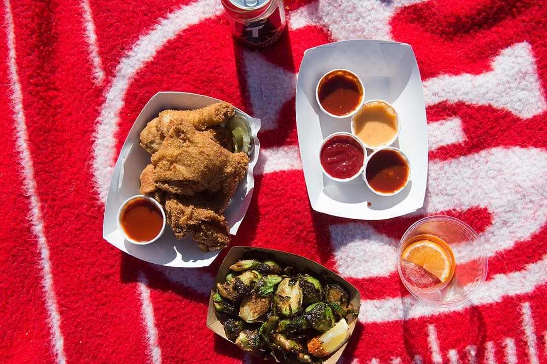 Overhead view of crispy fried chicken with dipping sauces, roasted Brussels sprouts, and drinks on a red patterned picnic blanket outdoors.