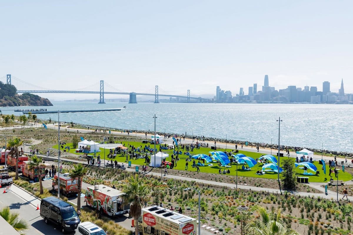 Aerial view of a waterfront park festival with tents, food trucks, and families gathered on the lawn, with the San Francisco skyline and Bay Bridge in the background.