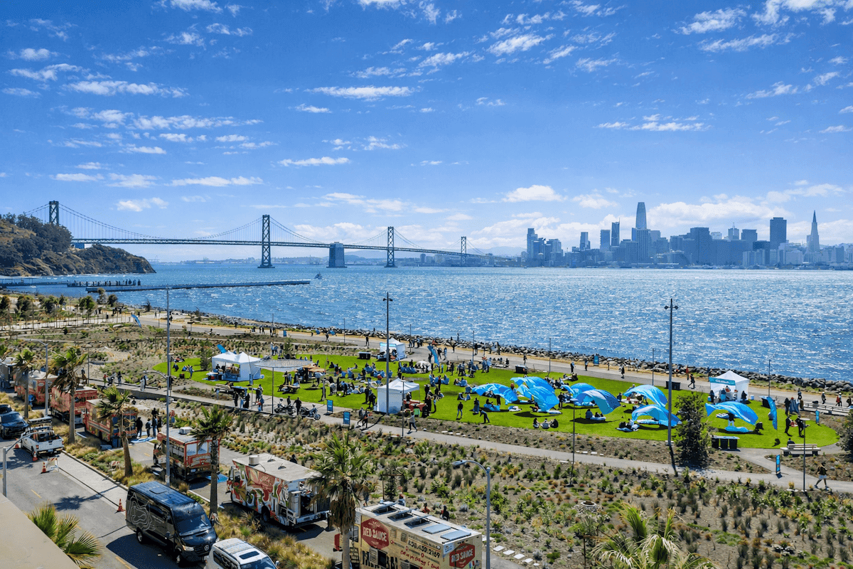 Waterfront park event with tents and food trucks along the San Francisco Bay, featuring views of the Bay Bridge and city skyline under a bright blue sky.