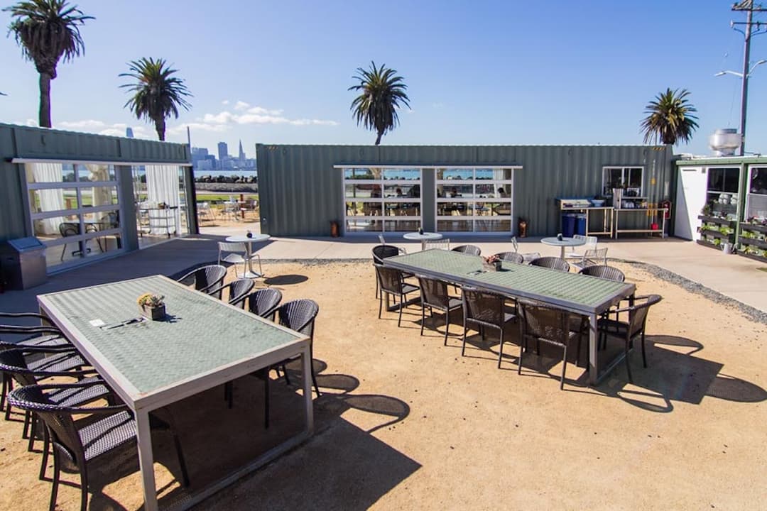Outdoor dining area made from converted shipping containers, with long communal tables and chairs set on a sandy patio lined with palm trees and views of the San Francisco skyline in the background.