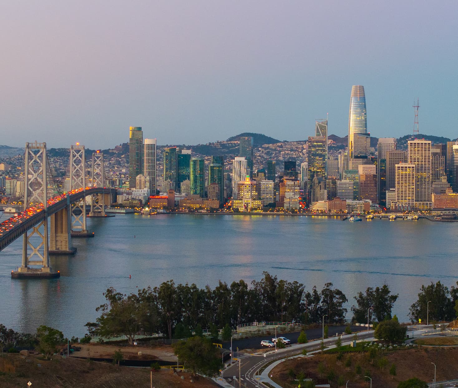 Evening view of the San Francisco skyline and Bay Bridge from Treasure Island, with city lights reflecting on the bay and waterfront roads in the foreground.