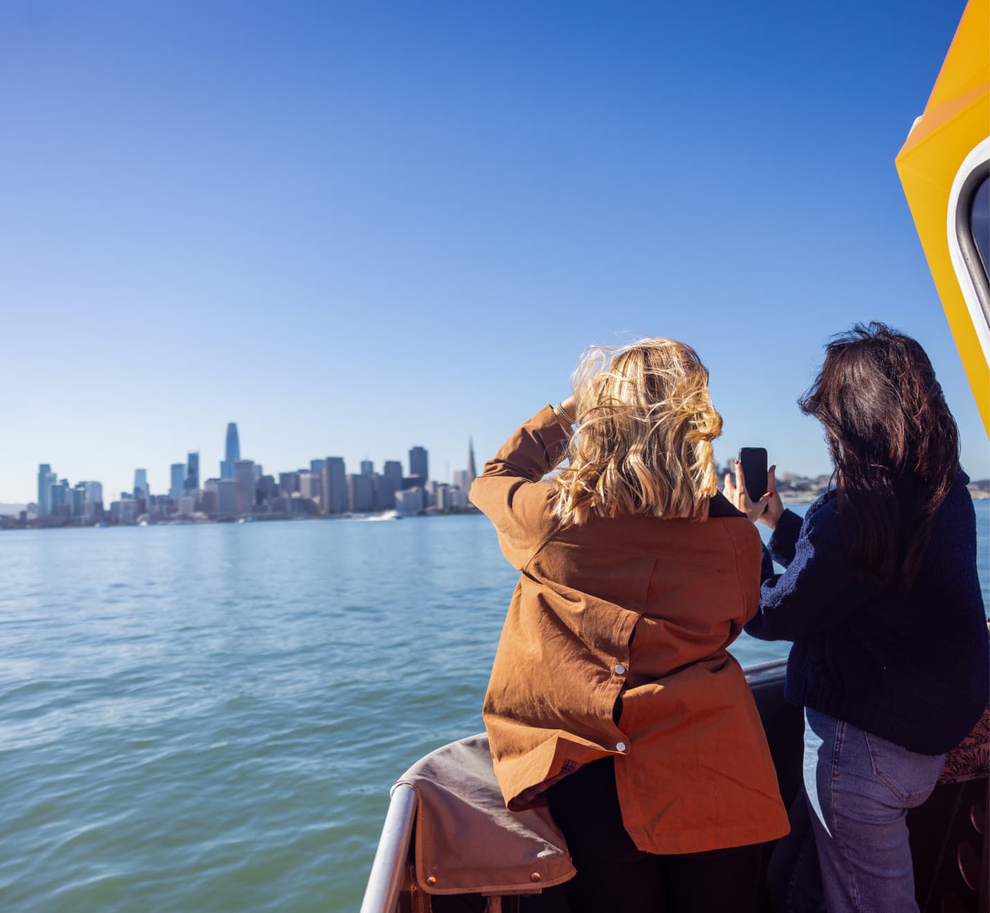 Two women standing on a ferry taking photos of the San Francisco skyline across the bay on a clear, sunny day.
