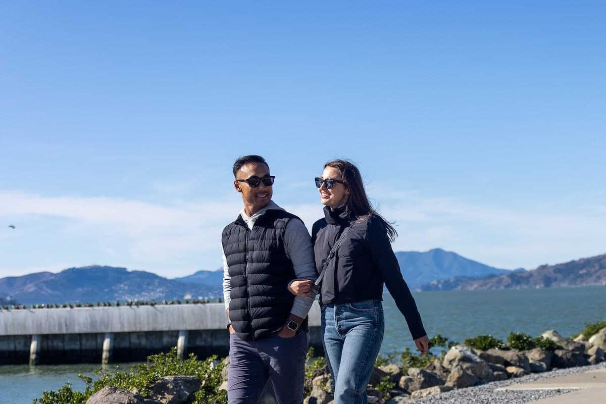 Couple wearing sunglasses walking hand in hand along a sunny waterfront path with rocky shoreline and mountains in the background.