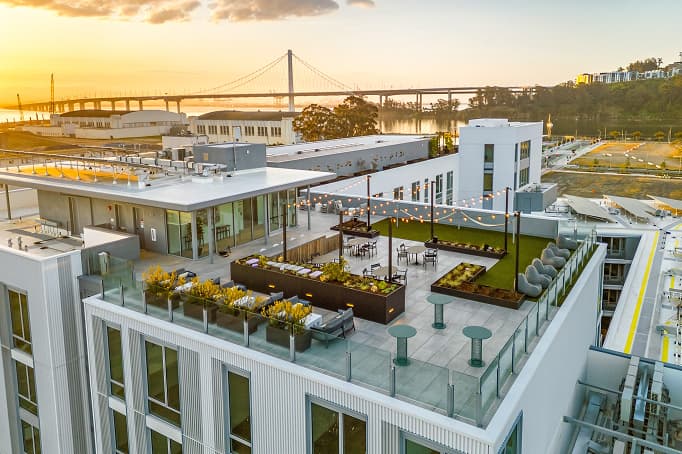 Rooftop terrace at a Treasure Island residential building with string lights, landscaped seating areas, and Bay Bridge views at sunset.