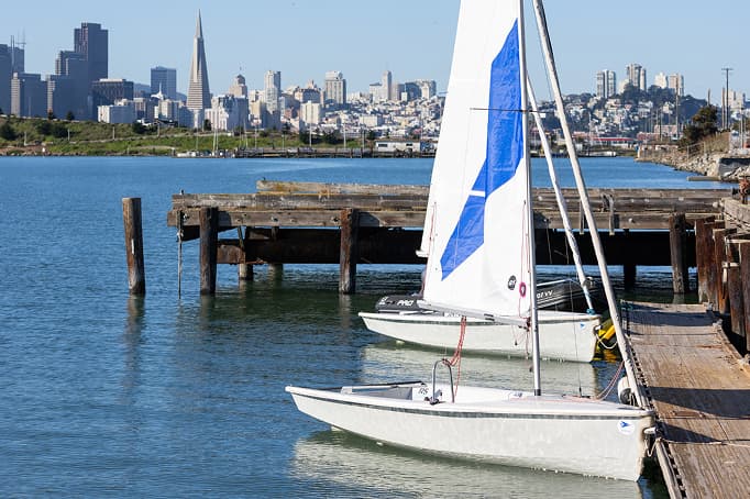Small sailboats docked at a wooden pier on calm bay waters, with the San Francisco skyline—including the Transamerica Pyramid—visible in the background under a clear sky.