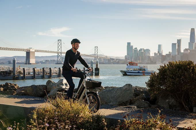 Cyclists riding along a paved waterfront bike path on Treasure Island, with a separated lane, open bay views, and the San Francisco skyline and Bay Bridge visible in the distance.