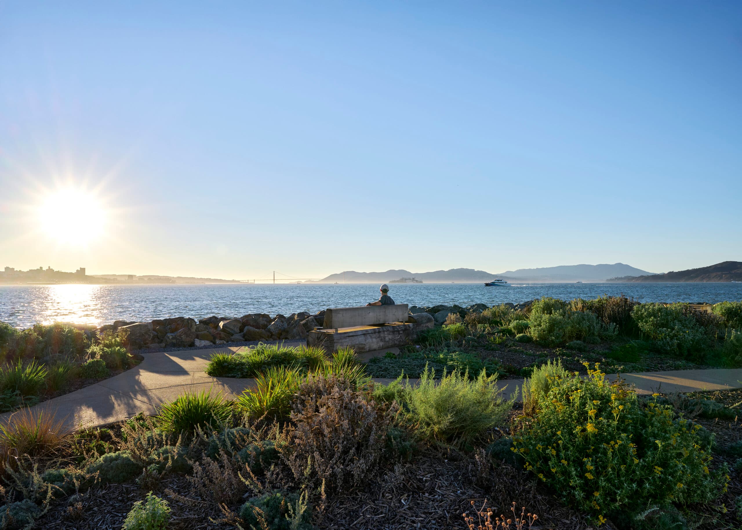 Sunlit bay view from a landscaped shoreline park with a person seated on a bench
