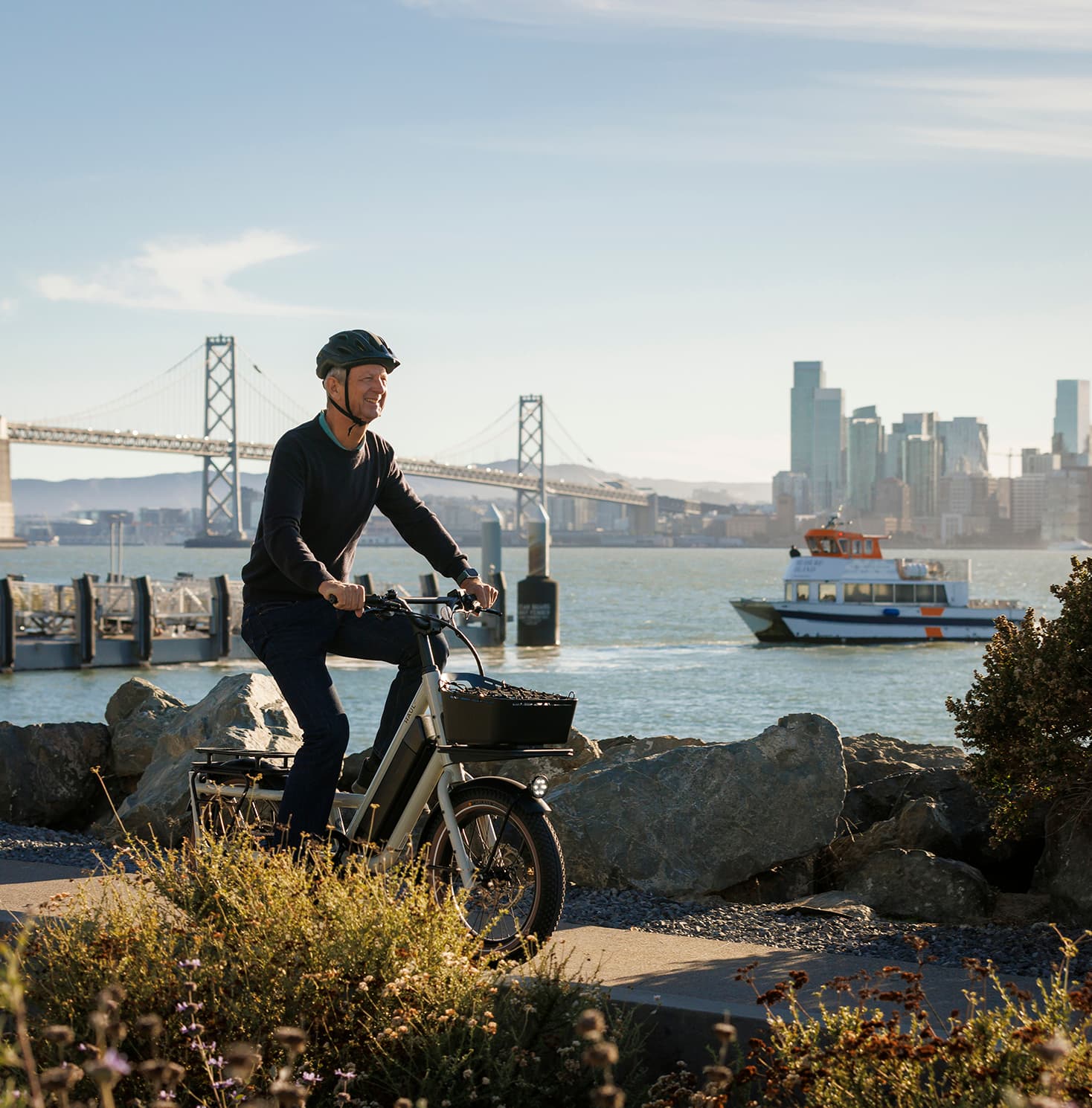 Person riding a bike along the Treasure Island waterfront with the Bay Bridge, San Francisco skyline, and a ferry in the background on a sunny day.
