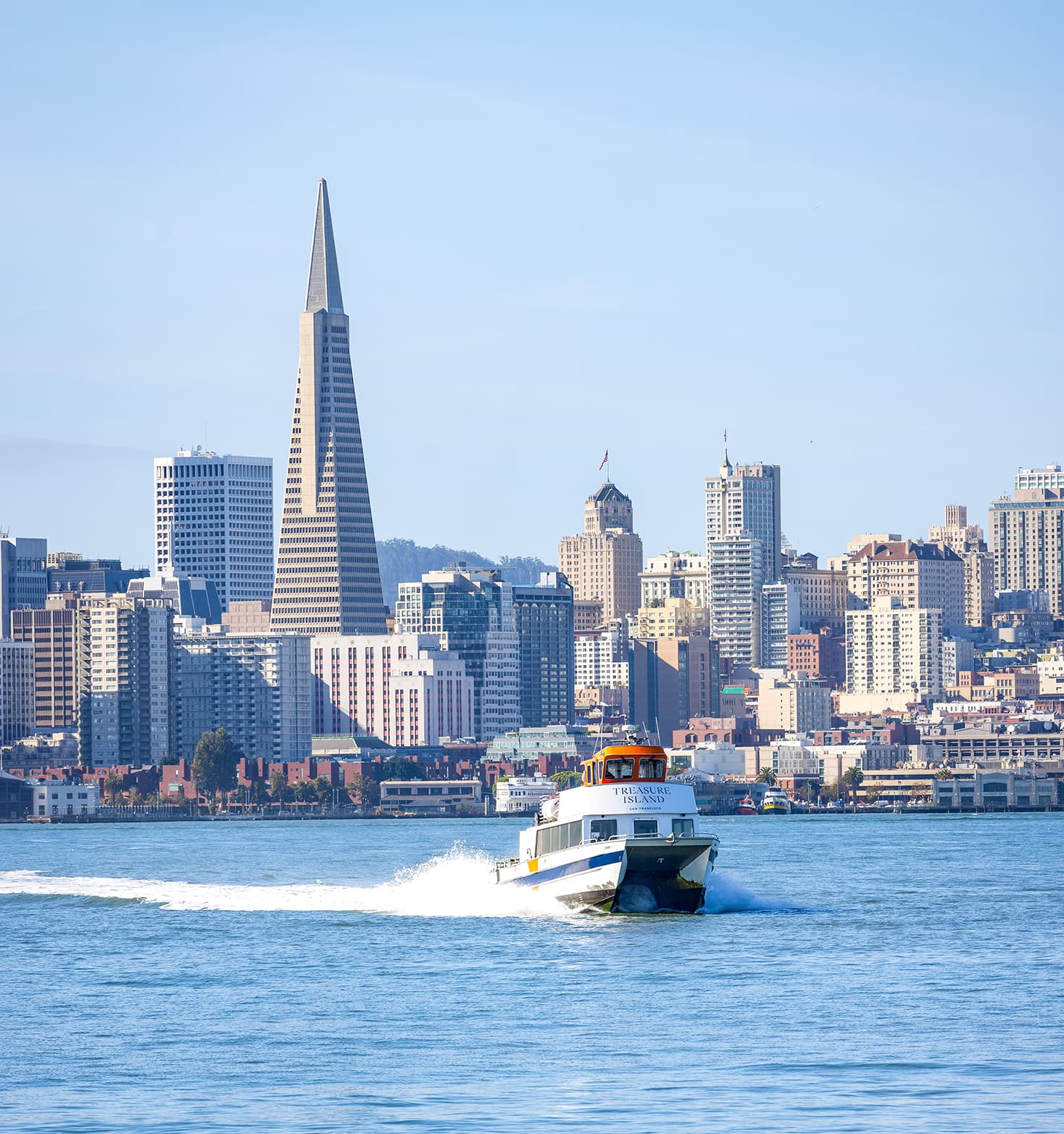 Treasure Island ferry speeding across the bay with the San Francisco skyline and Transamerica Pyramid in the background on a clear day.