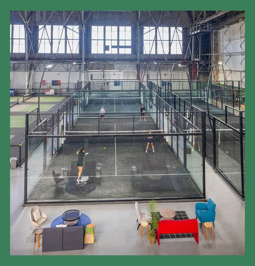 People playing padel on enclosed courts at a Bay Padel event on Treasure Island, with multiple courts, spectators nearby, and a large indoor venue with high ceilings and lighting overhead.
