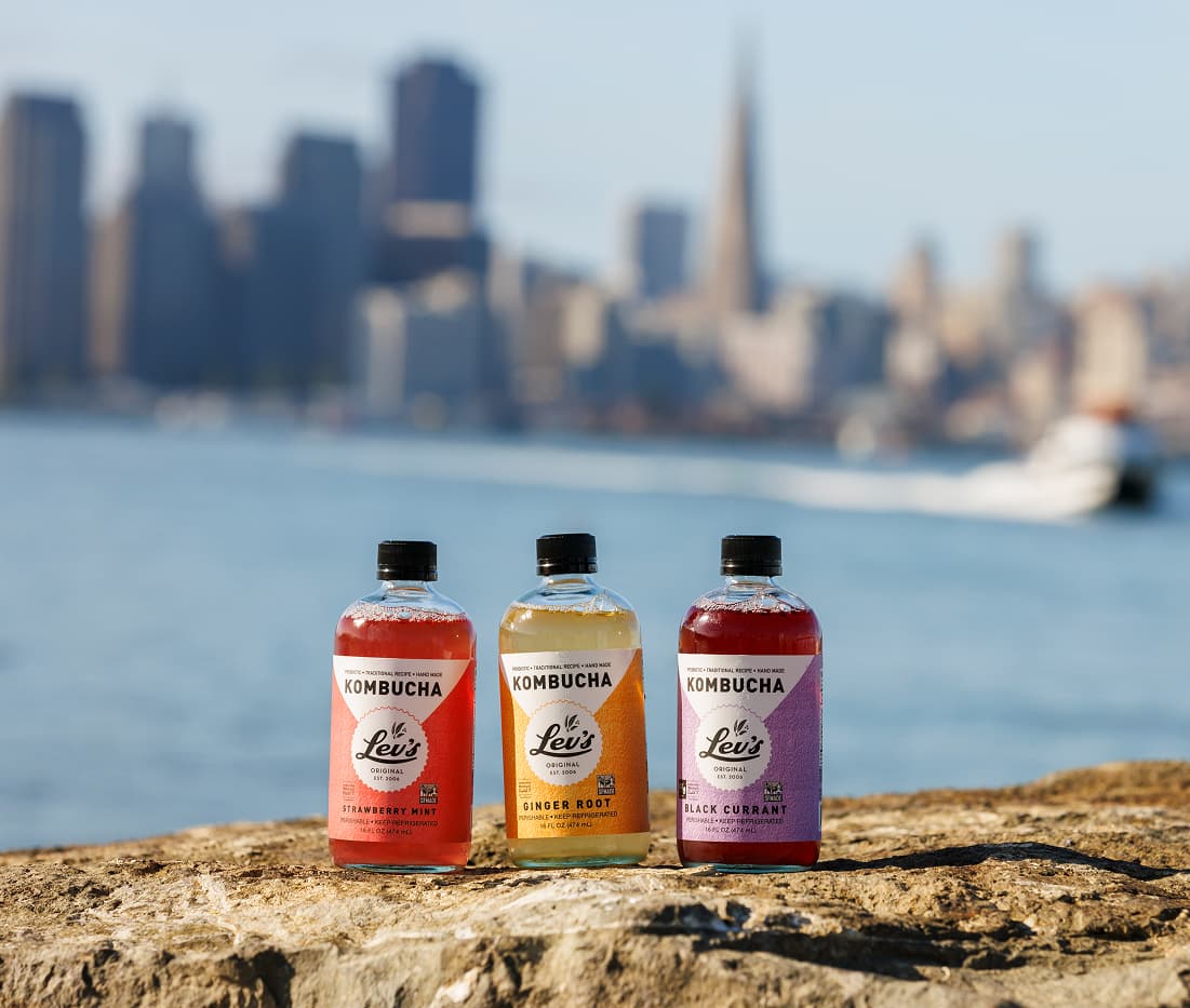 Three bottles of Lev’s Original kombucha in Ginger Root and Raspberry Mint flavors displayed waterfront with 2015 and 2017 Good Food Awards medals, highlighting award-winning probiotic craft beverages.