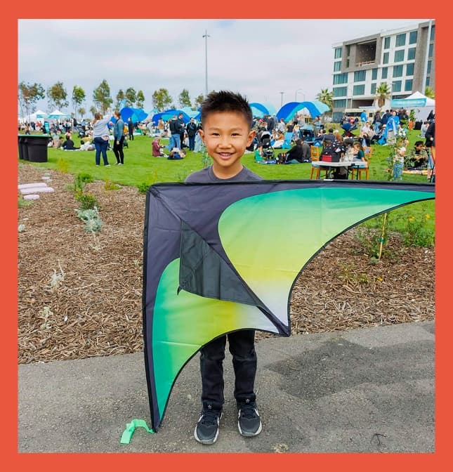 Smiling child holding a green-and-black kite at a busy waterfront park event