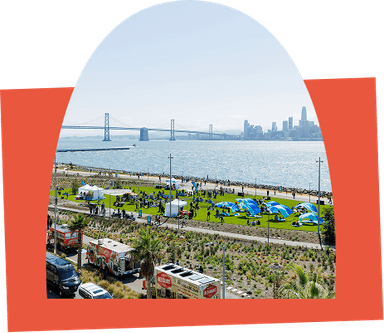 Aerial view of a waterfront park with people on the lawn, food trucks, and the Bay Bridge and city skyline in the background