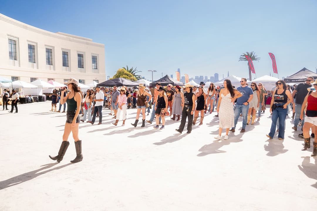 Large group of people line dancing outdoors at a sunny festival with vendor tents and city skyline in the background