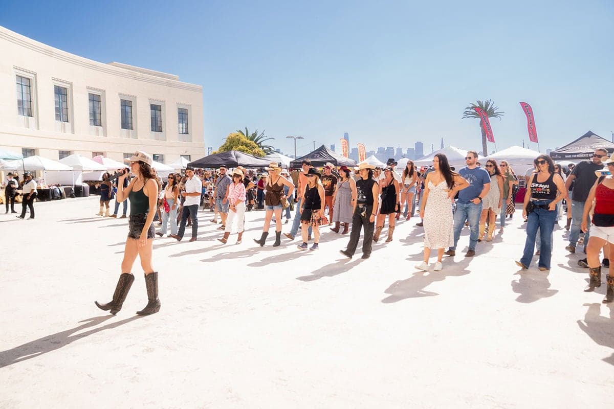 Large group of people line dancing outdoors at a sunny festival with vendor tents and city skyline in the background