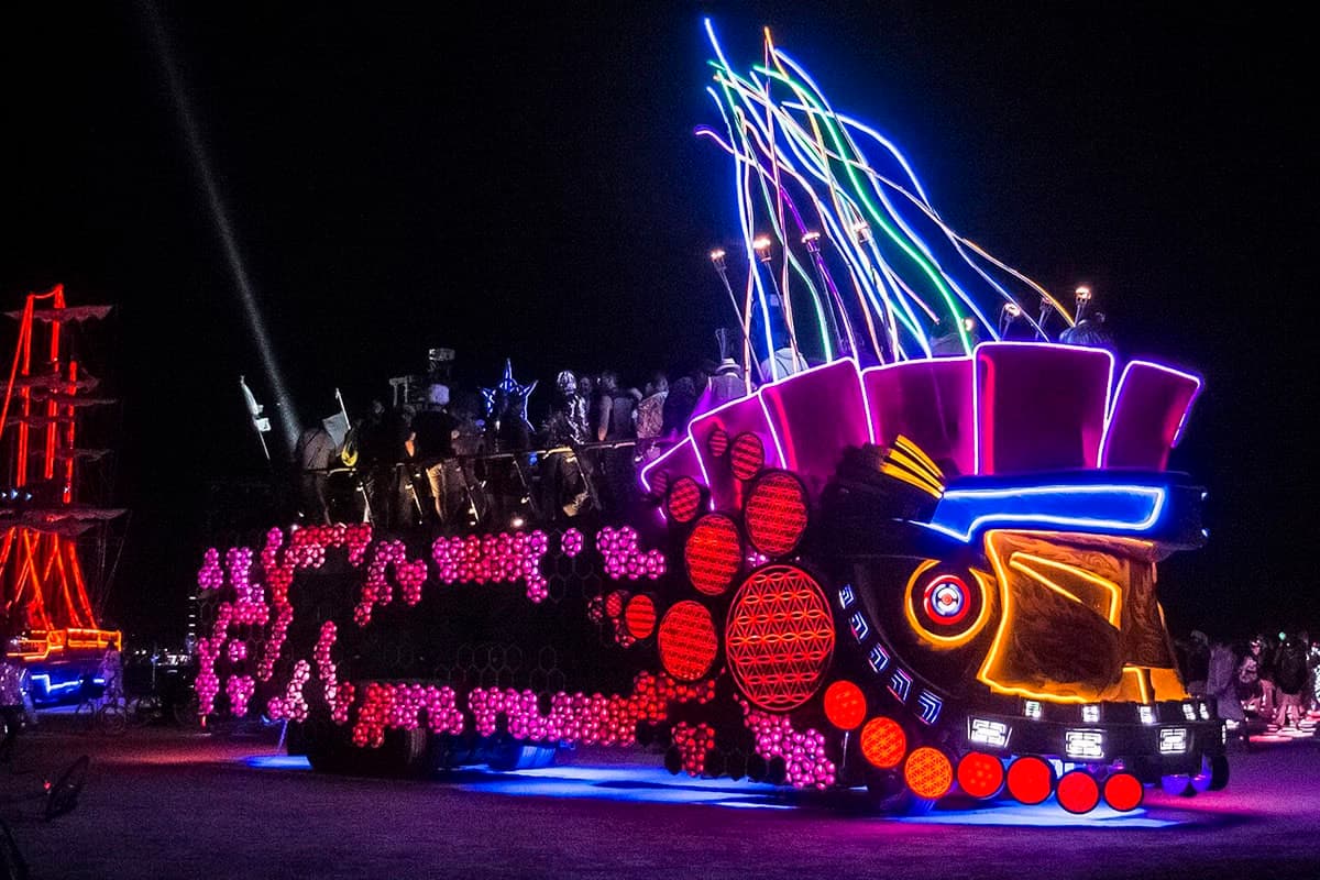 Colorful illuminated art car with neon lights and performers at a nighttime festival or desert event