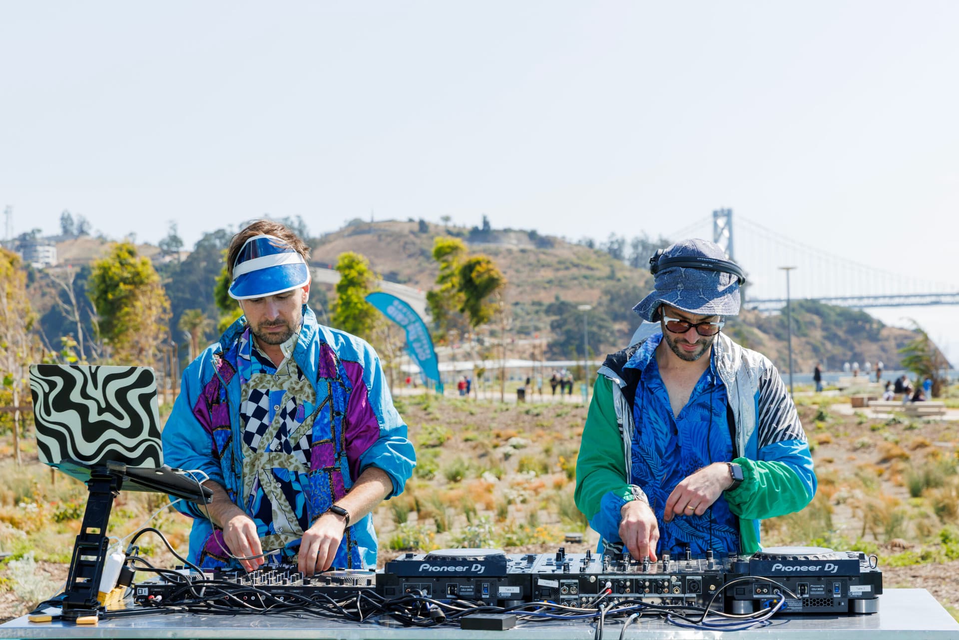 Two DJs mixing music on a Pioneer DJ setup at an outdoor waterfront location with hills and bridge in background