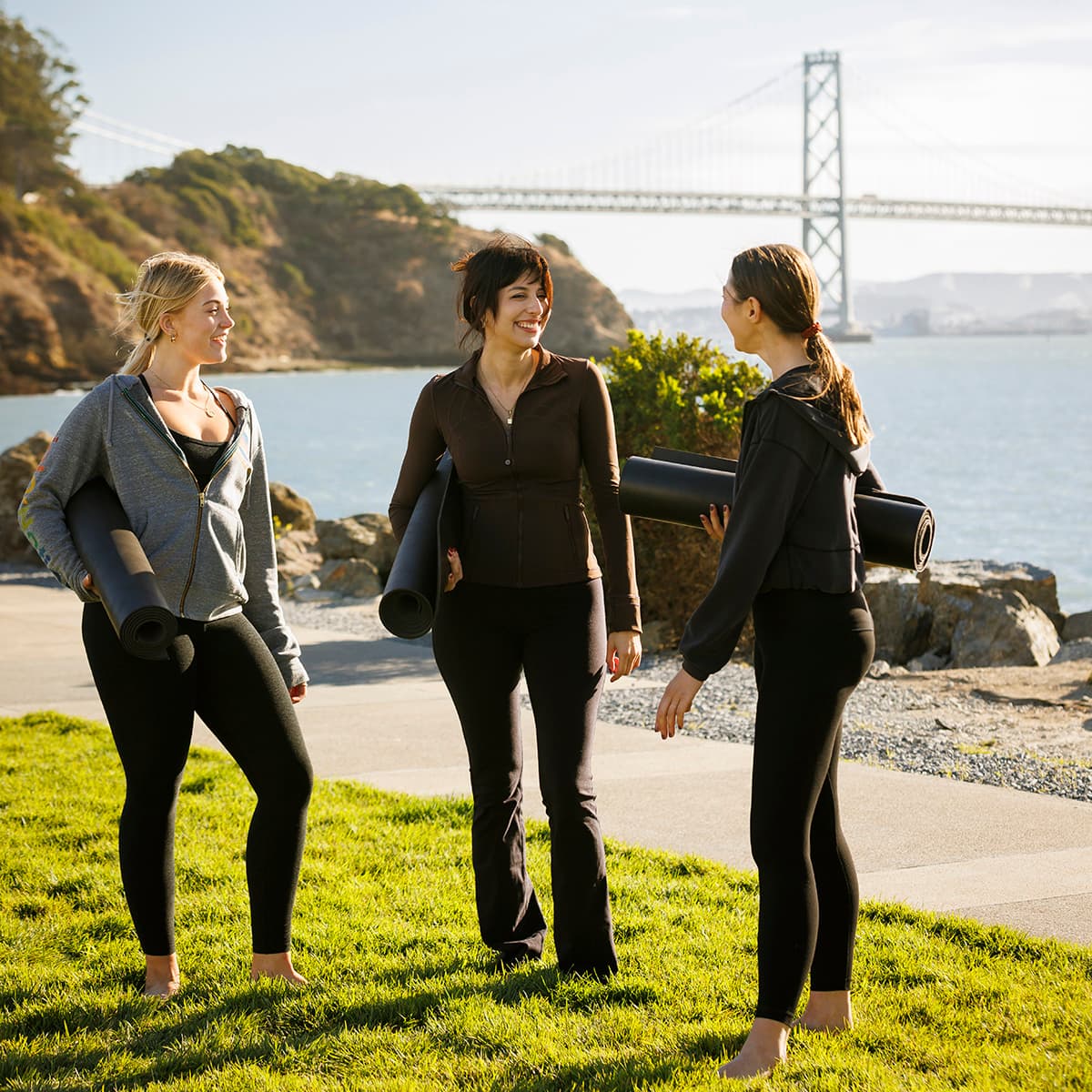 Three people holding yoga mats and talking by the waterfront, with the Bay Bridge in the background.