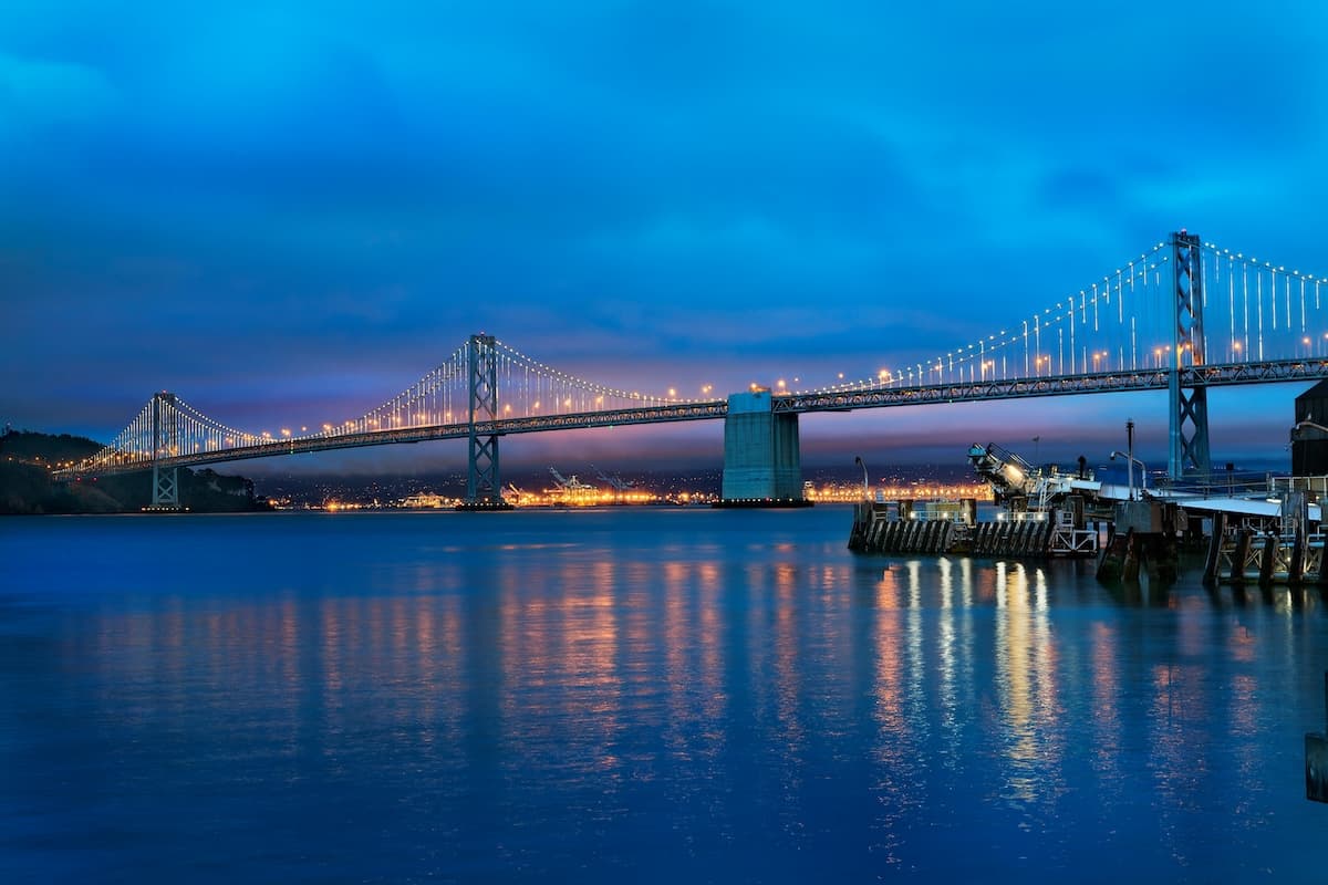 A long suspension bridge illuminated with lights stretches across calm water at dusk, with a deep blue sky and reflections shimmering on the surface; a small pier with equipment sits in the foreground on the right.