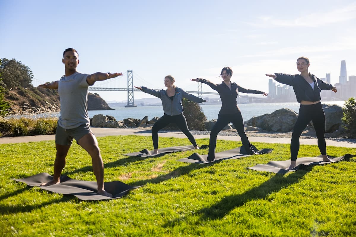 Four people practice yoga on mats in a grassy waterfront park, standing in a warrior pose with arms extended. Behind them, a large suspension bridge and a distant city skyline rise across the water on a bright, sunny day.
