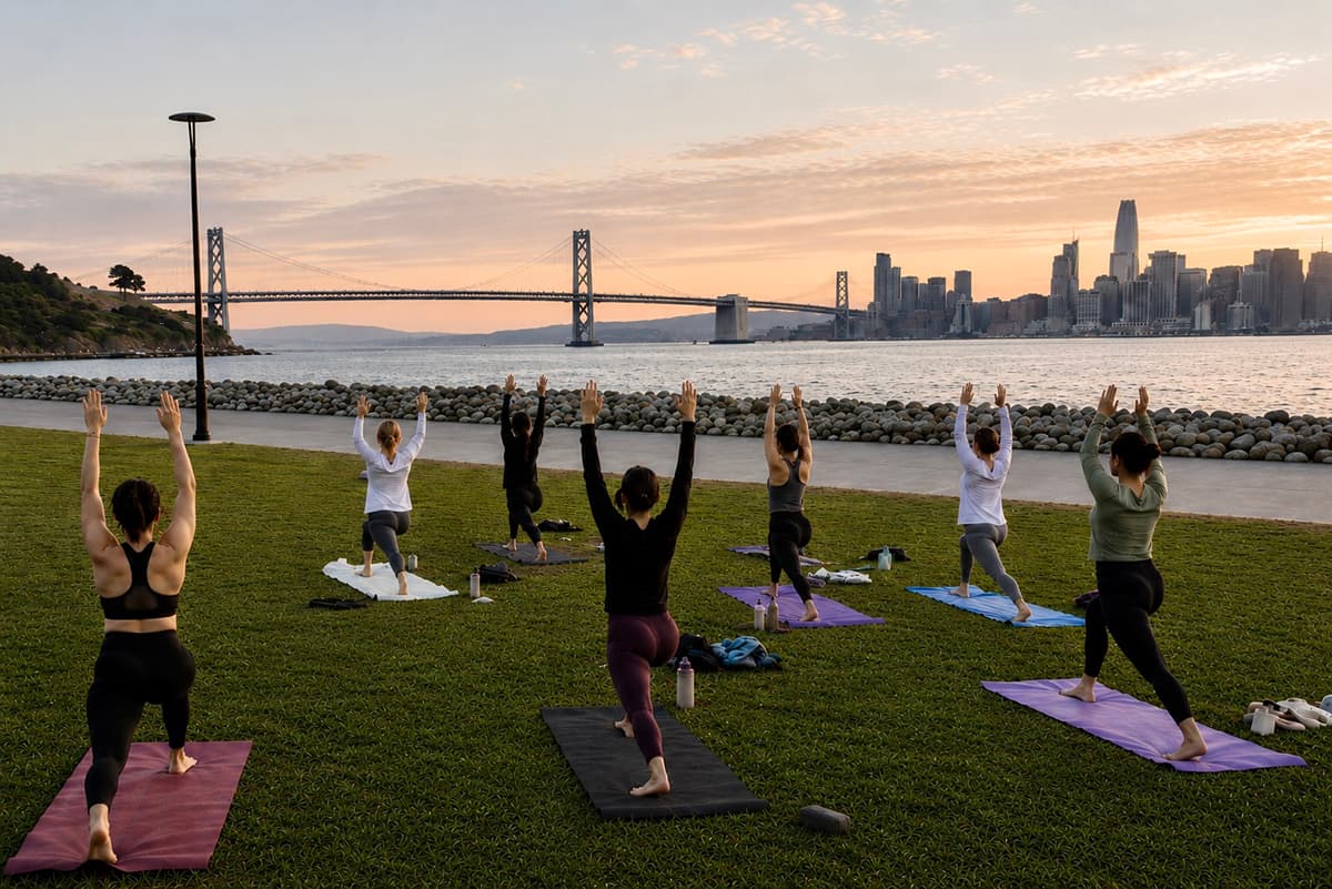 People practicing sunset yoga on a waterfront lawn at Treasure Island with the Bay Bridge and San Francisco skyline in the background.