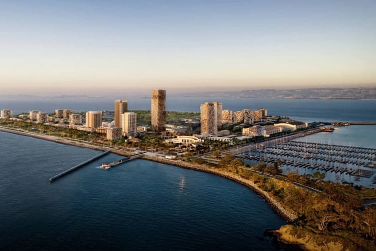 Aerial view of a modern waterfront city skyline with high-rise residential towers, marina harbor, and coastal promenade at sunset.