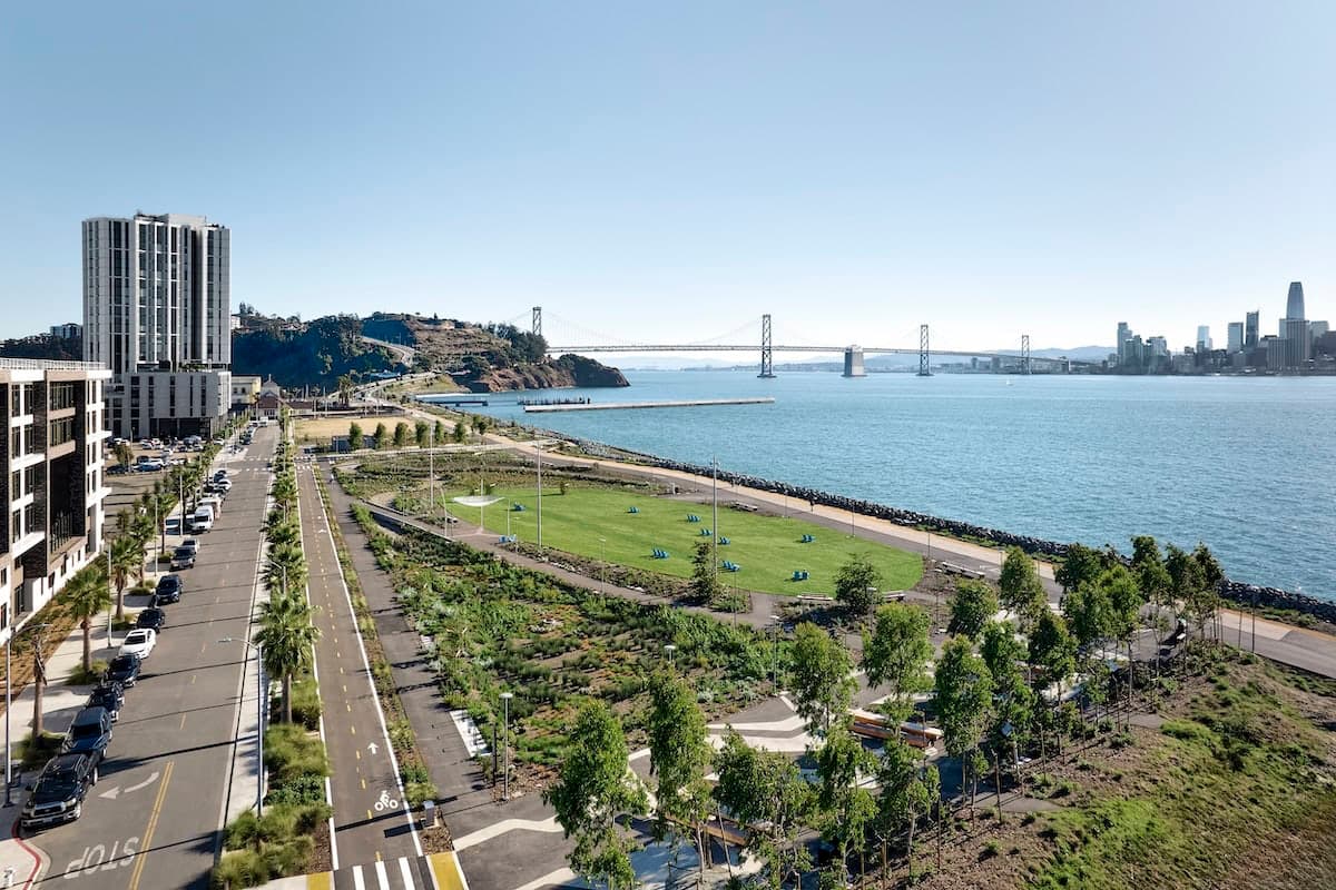 Aerial view of a waterfront park and residential buildings along the San Francisco Bay with the Bay Bridge and city skyline in the distance.