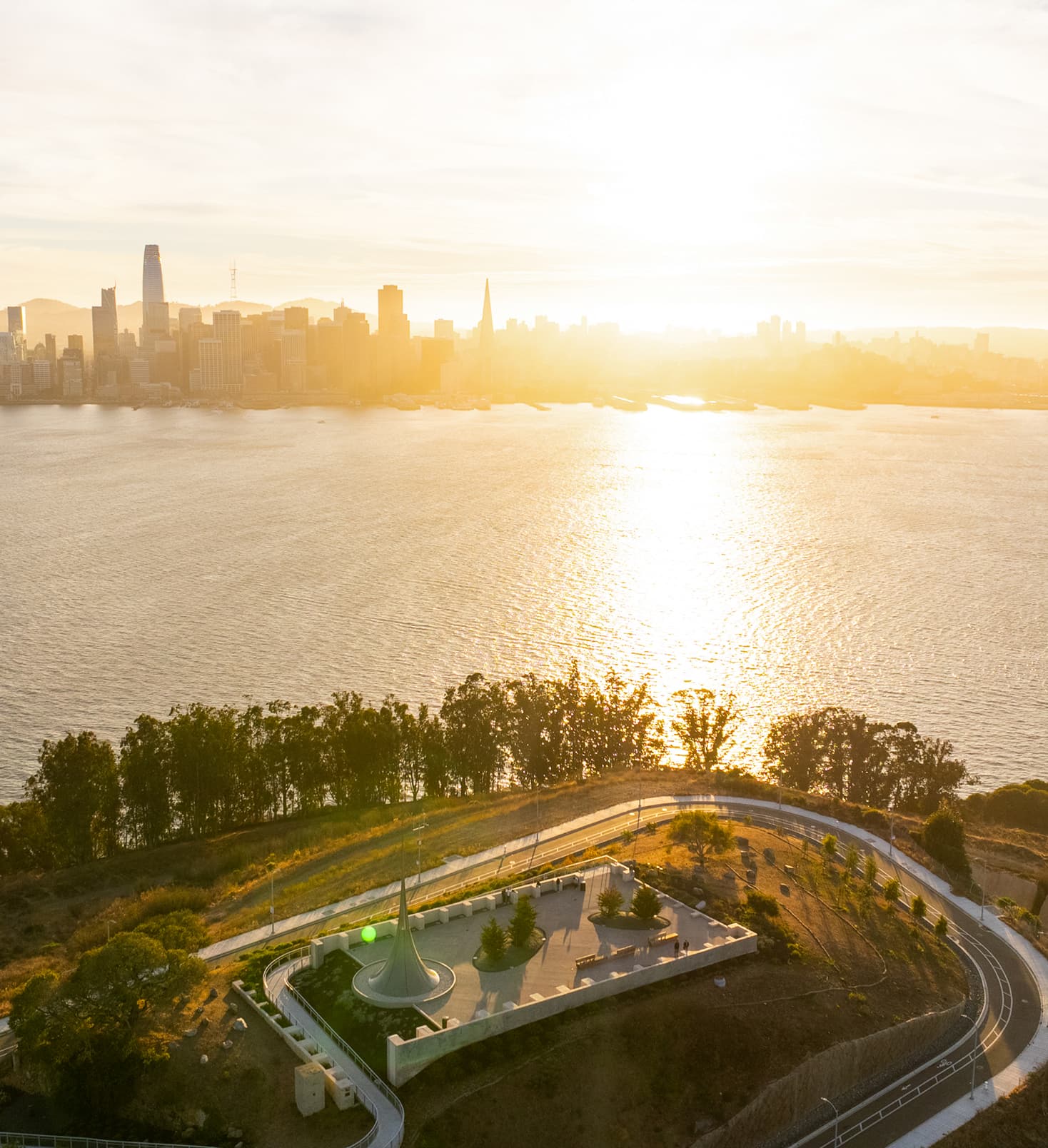 Aerial sunset view over the San Francisco skyline and bay, with golden light reflecting across the water. In the foreground, a landscaped overlook on Treasure Island features a sculptural spire, terrace seating, and winding pathways along the hillside.