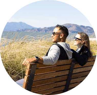 A couple sits side by side on a wooden bench overlooking the water, surrounded by tall coastal grasses. Both wear sunglasses and appear relaxed as they take in the view of the bay and distant hills under clear skies.