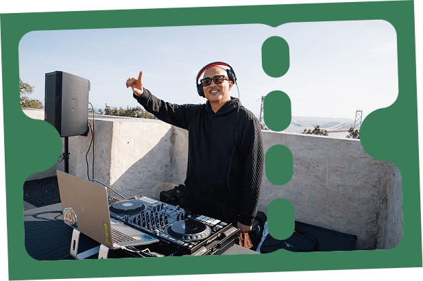 DJ performing on a rooftop terrace with turntables and laptop, wearing headphones and pointing outward, with the Bay Bridge visible in the background during a daytime outdoor event.