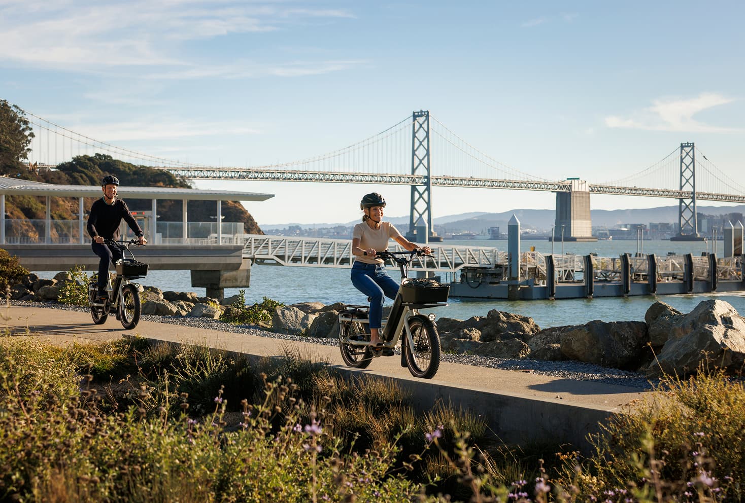 Two cyclists riding electric bikes along a waterfront path near the San Francisco–Oakland Bay Bridge, with marina docks and bay views in the background on a clear day.