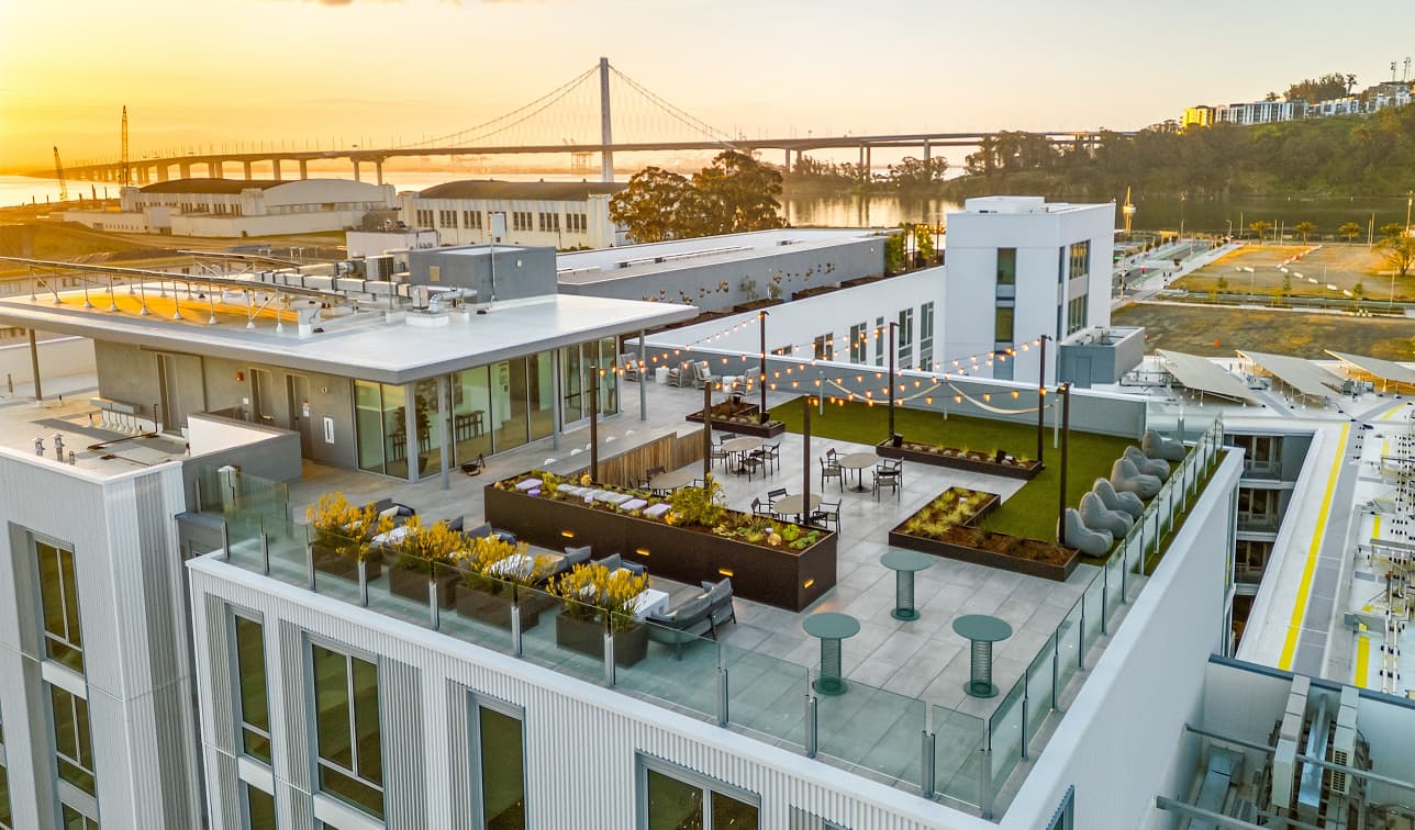 Rooftop terrace at Hawkins featuring modern outdoor seating, landscaped planters, string lights, and expansive sunset views of the Bay Bridge and San Francisco Bay on Treasure Island.