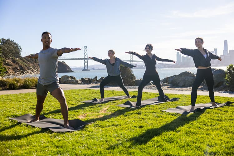 People practicing yoga on mats by the waterfront, with the Bay Bridge and San Francisco skyline in the background.