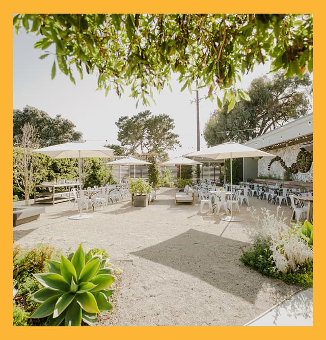 Sunny outdoor patio at Aracely Café on Treasure Island featuring garden landscaping, shaded umbrellas, communal tables, and mural art for al fresco dining in San Francisco.