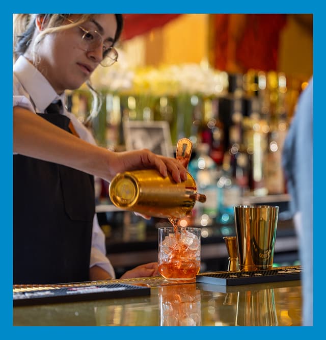 Bartender pouring a cocktail over ice at Gold Bar, with bottles, bar tools, and warm ambient lighting in the background.