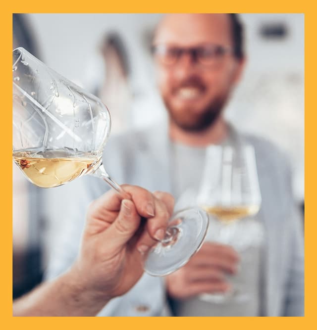Two people tasting white wine indoors, with one person swirling a glass in the foreground while another smiles in the background holding their own glass, suggesting a relaxed winery or tasting room setting.