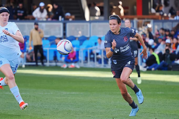 A women’s soccer player in a dark Bay FC uniform sprinting toward the ball during a match, with an opposing player in a light kit nearby and spectators visible in the background.