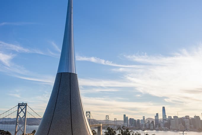 A tall, sleek sculptural spire rising in the foreground at a waterfront park, with the San Francisco skyline and the Bay Bridge stretching across the bay under a clear sky.