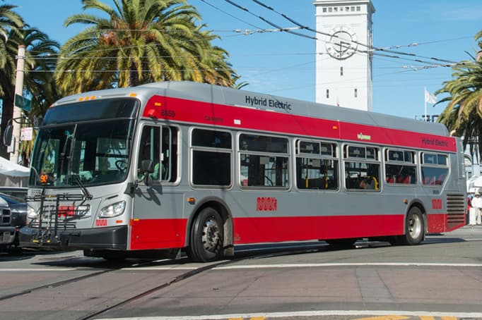 Red and silver SFMTA Muni 25 Treasure Island bus driving through San Francisco streets with the Ferry Building clock tower in the background.