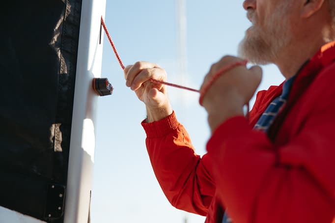 Close-up of a man in a red jacket adjusting a rope on a boat or nautical structure, focusing on the knot and hardware against a clear sky.