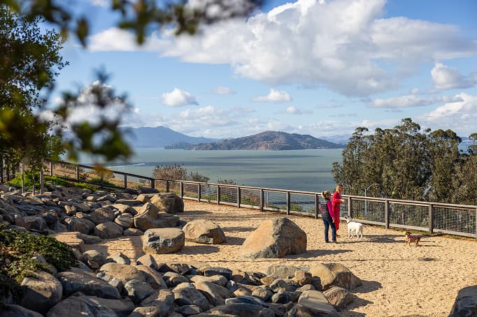 A scenic dog park overlooking the bay, with large rocks, a fenced gravel area, and a person standing with two dogs against a backdrop of water and distant hills under a partly cloudy sky.