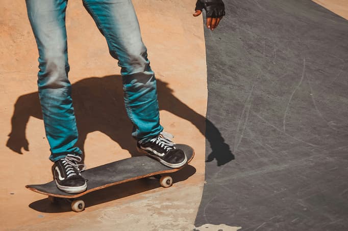 A close-up of a skateboarder riding along the curved edge of a skatepark ramp, wearing jeans, black sneakers, and a glove, with the board rolling along the smooth concrete surface.