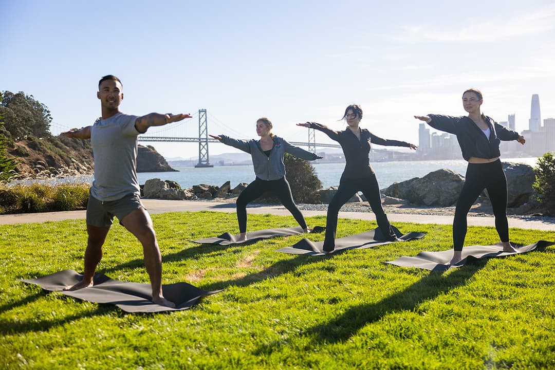 People practicing yoga on mats by the waterfront, with the Bay Bridge and San Francisco skyline in the background.