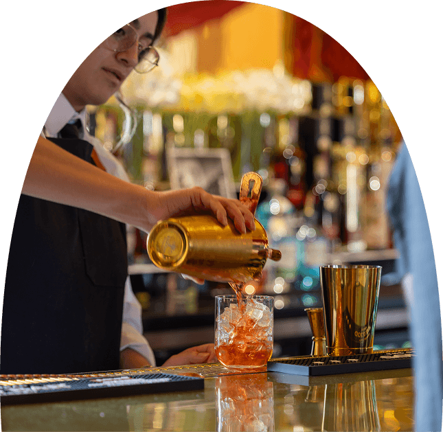 Bartender pouring a cocktail over ice at Gold Bar, with bottles, bar tools, and warm ambient lighting in the background.