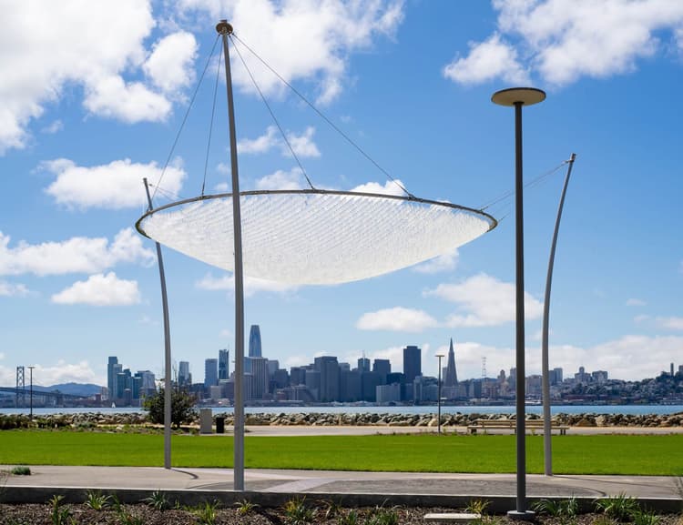 Kinetic sculpture called ‘Canopy of Sky’ suspended above a public park walkway on Treasure Island, with thousands of small translucent discs hanging from cables and shimmering in the wind against views of San Francisco Bay and skyline.