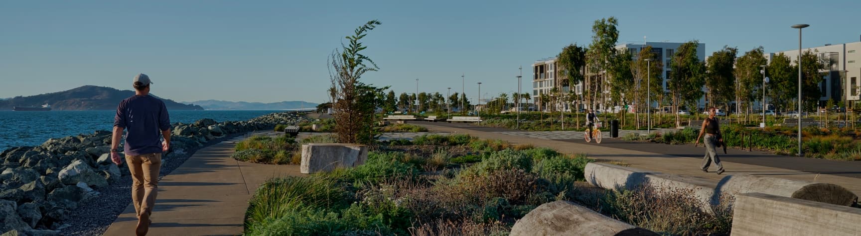 People walking and sitting along a waterfront promenade on Treasure Island, with a paved path, rocky shoreline, and wide views of San Francisco Bay under a clear sky.