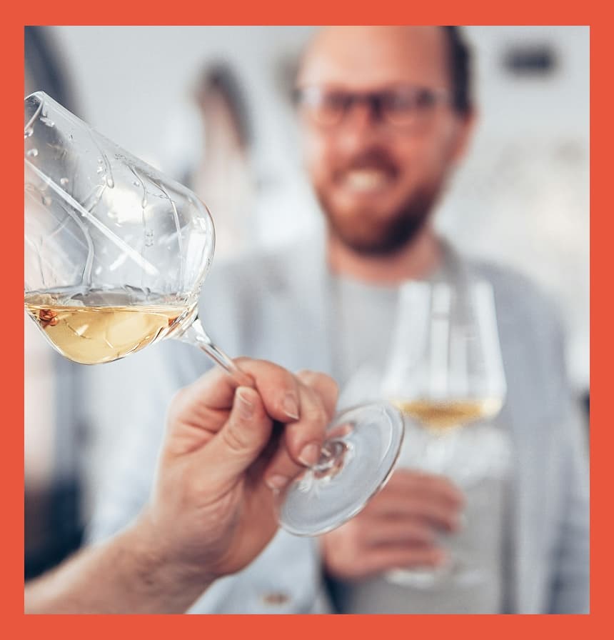 Two people tasting white wine indoors, with one person swirling a glass in the foreground while another smiles in the background holding their own glass, suggesting a relaxed winery or tasting room setting.